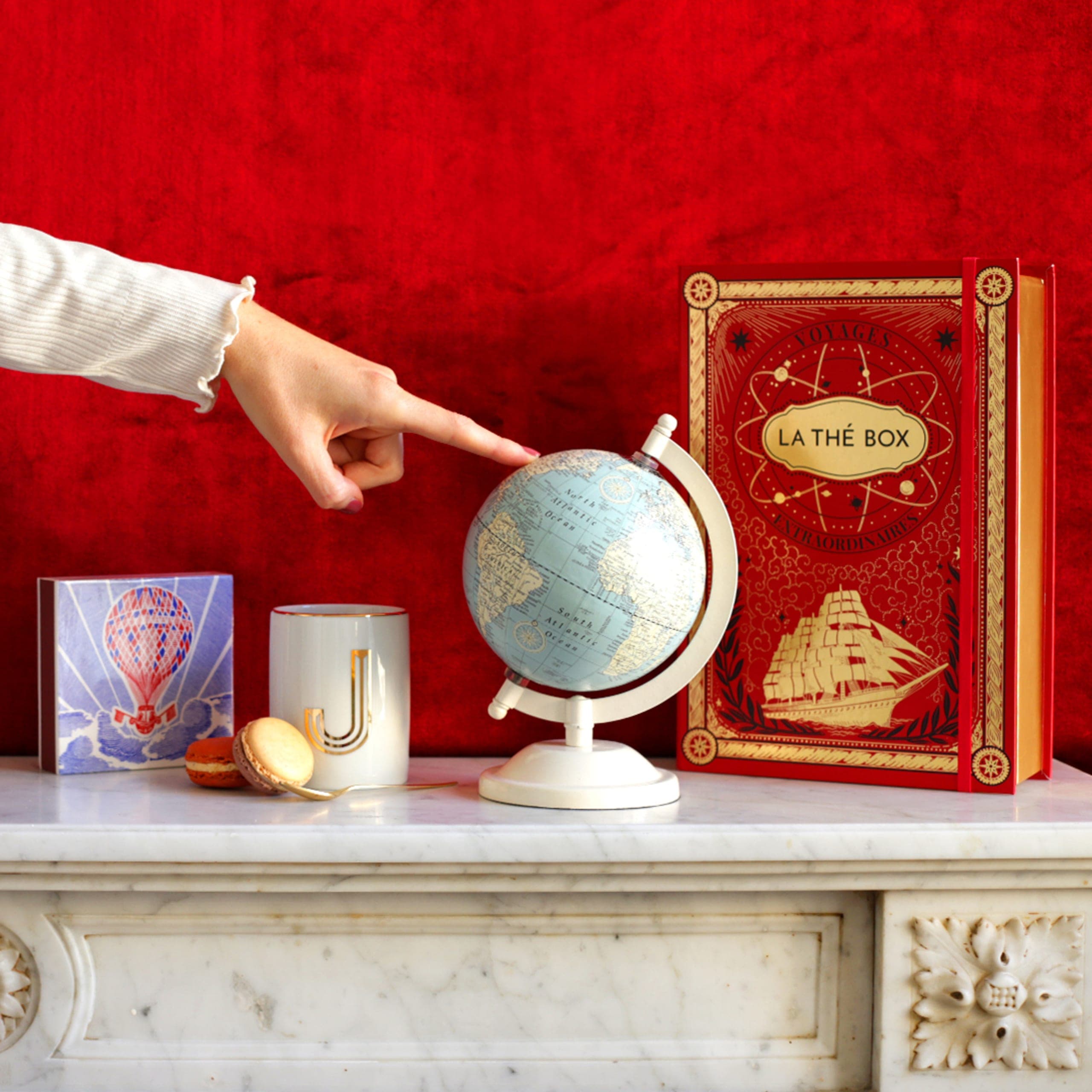 Red book-shaped box with gold-framed sailing ship engraving and ornamental borders, displayed upright on marble surface.